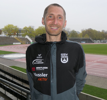 lächelnder Mann in Sportjacke mit SSV 1846 Schriftzug vor Leichtathletikbahn
